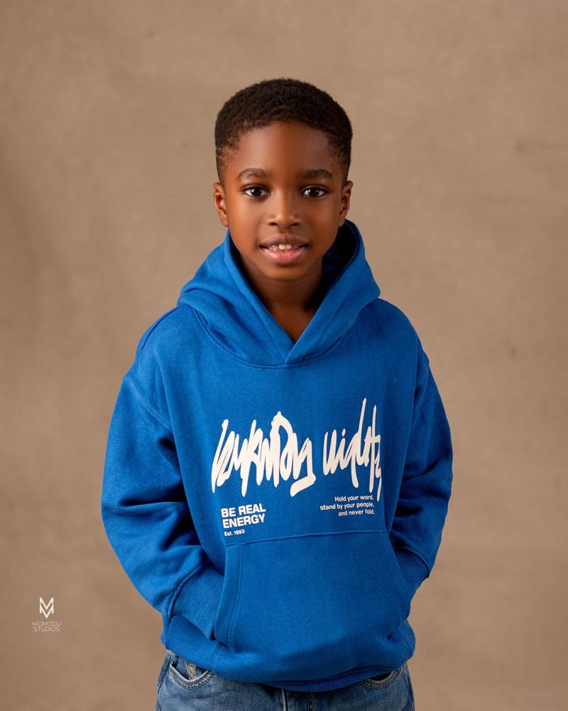 A young boy in a blue hoodie smiling at the camera against a tan backdrop