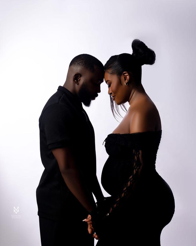 An expecting couple in black, foreheads touching, against a pale grey backdrop
