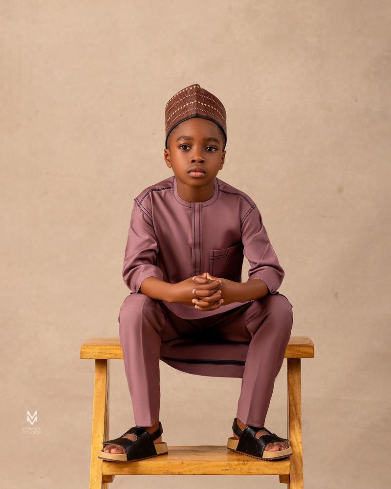 A boy in a maroon kaftan and cap seated on a wooden stool