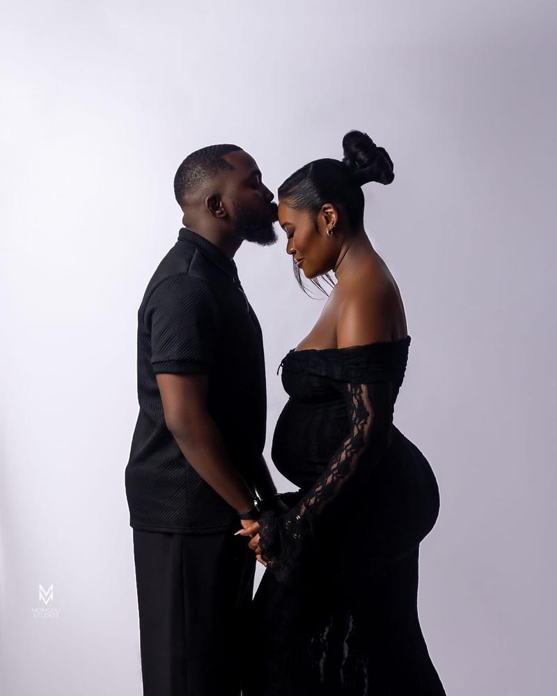 A man kisses his pregnant partner's forehead, both dressed in black