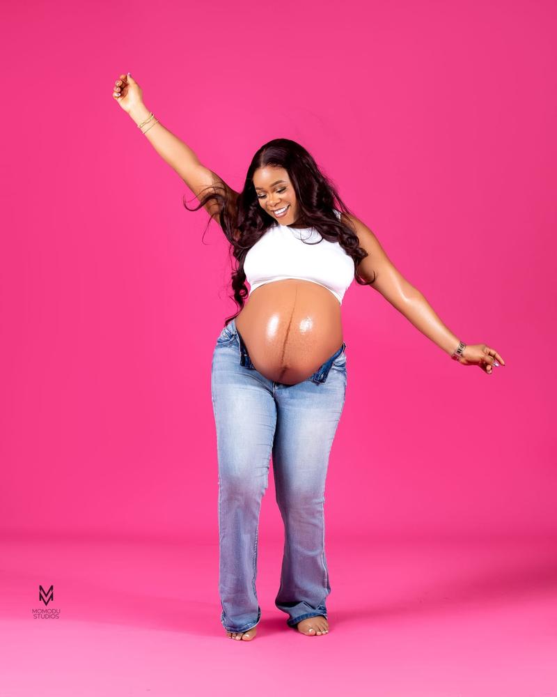 A pregnant woman in a white crop top and jeans laughing with one arm raised, pink backdrop