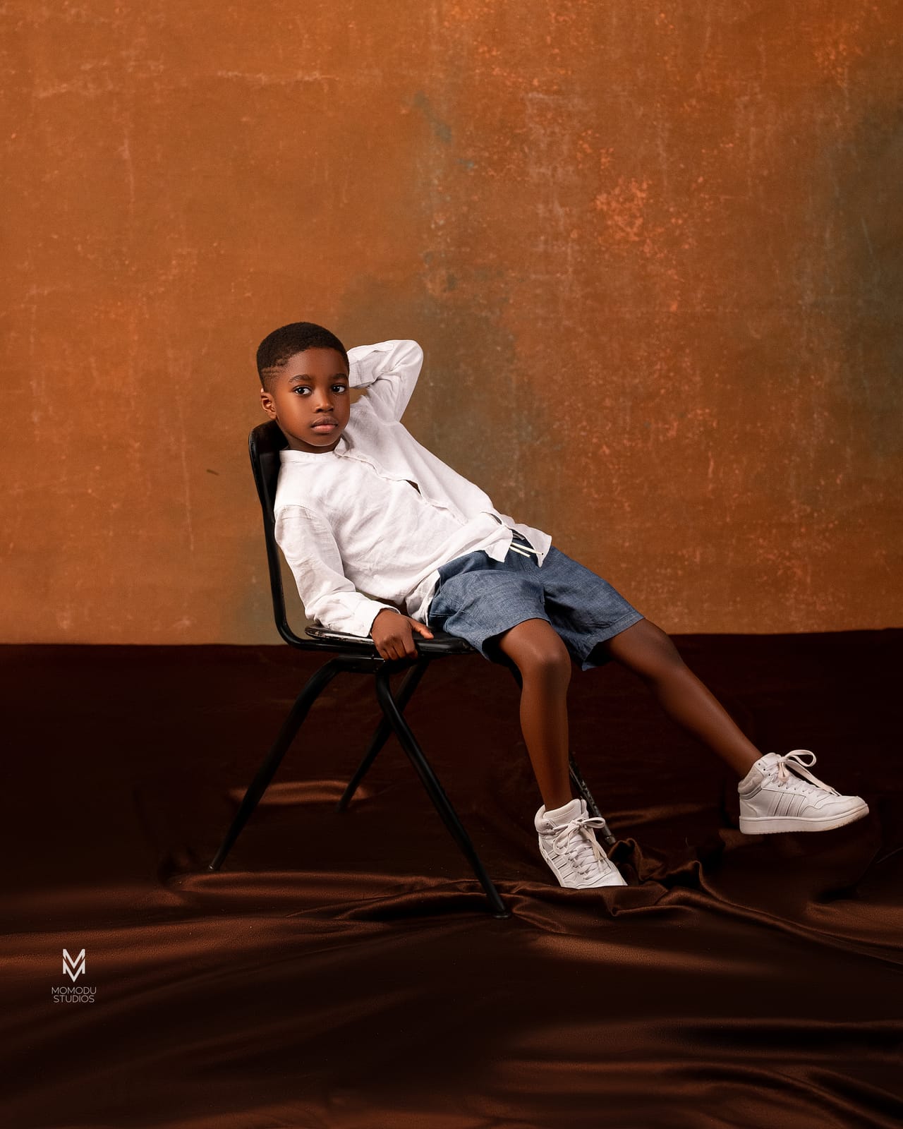 A boy in a white shirt and denim shorts lounging on a director's chair, brown backdrop