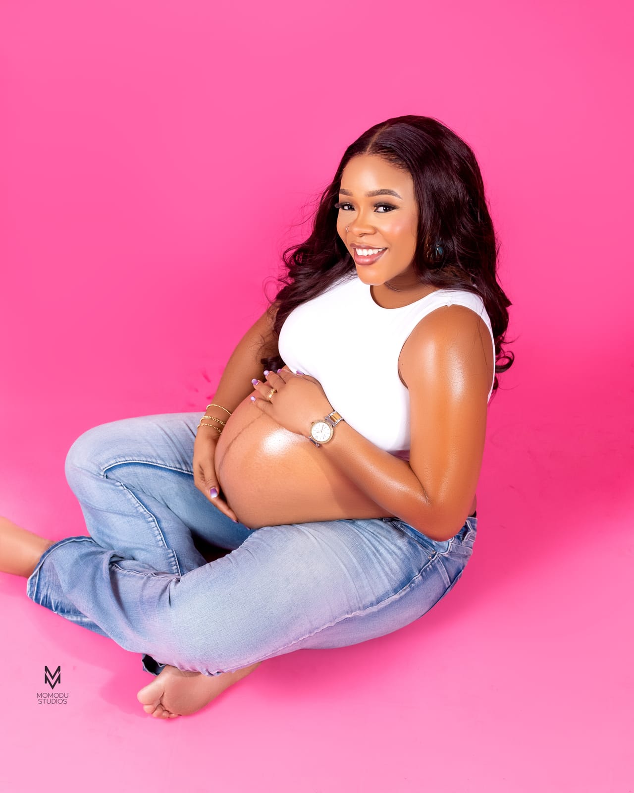 A pregnant woman in a white top and jeans sitting cross-legged on a pink set