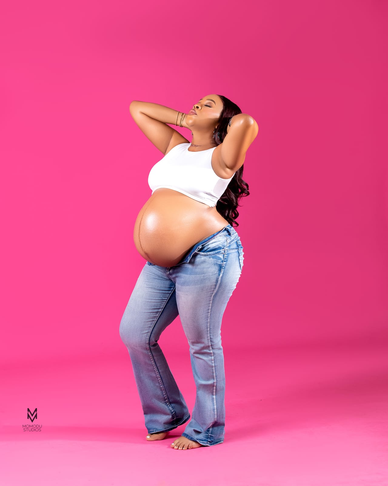 A pregnant woman in a white crop top posing with her hands in her hair, pink backdrop