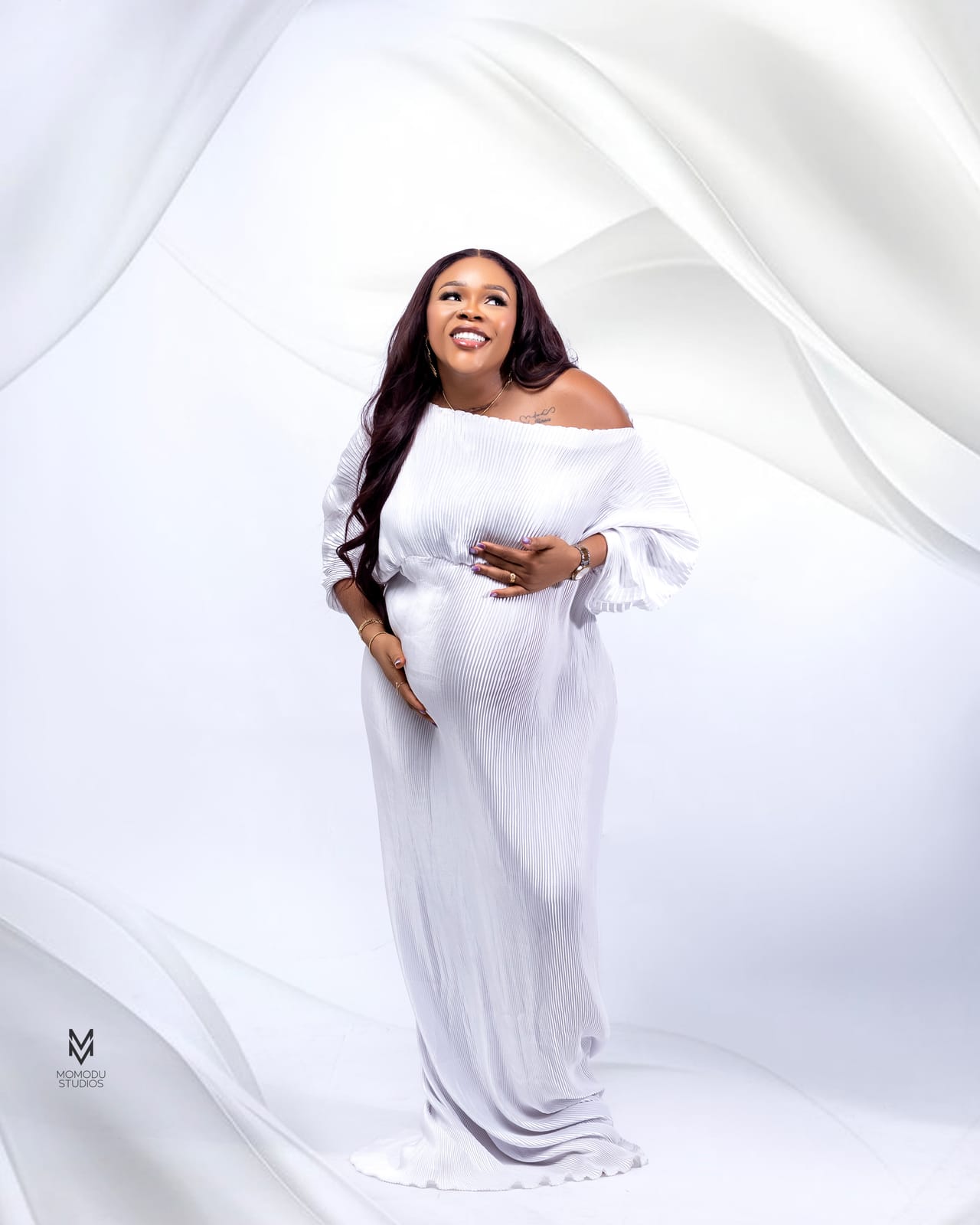 A pregnant woman in a flowing white off-shoulder gown against white drapes