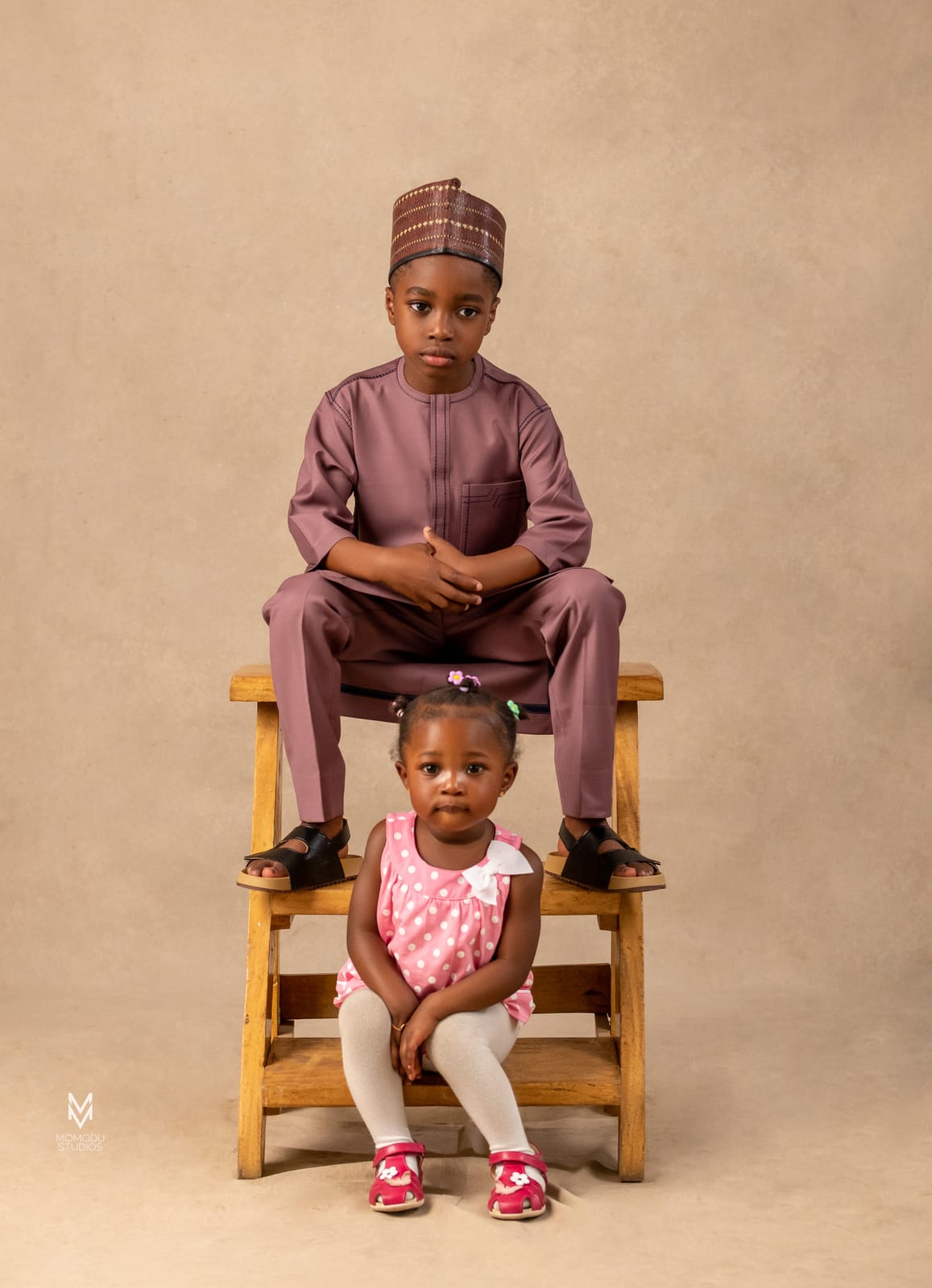 A boy in a maroon kaftan seated on a stool with his little sister below him