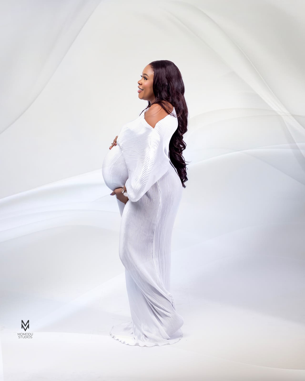 A pregnant woman in a white gown photographed in profile against white drapes