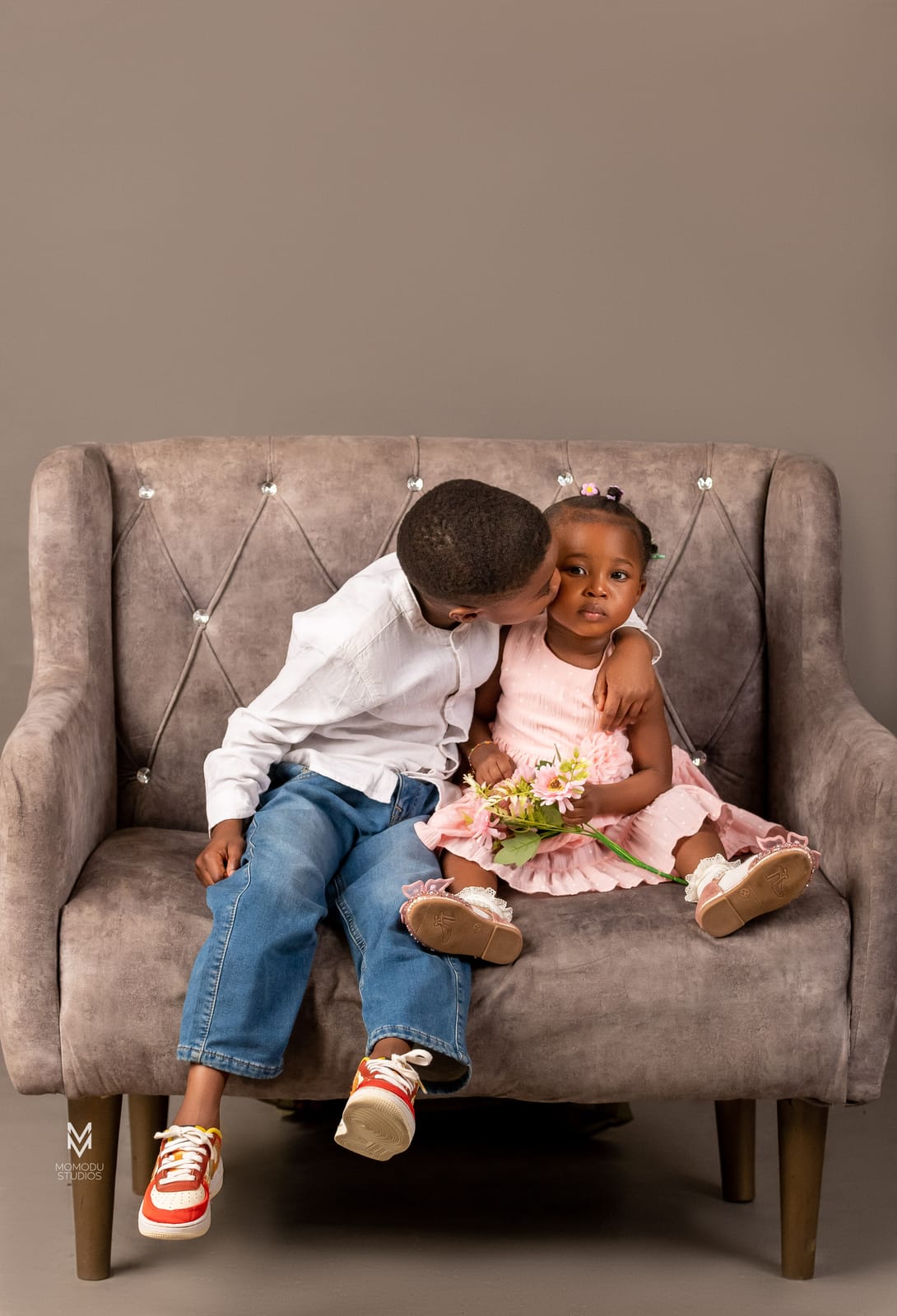 A boy kisses his baby sister's cheek as they sit on a grey sofa