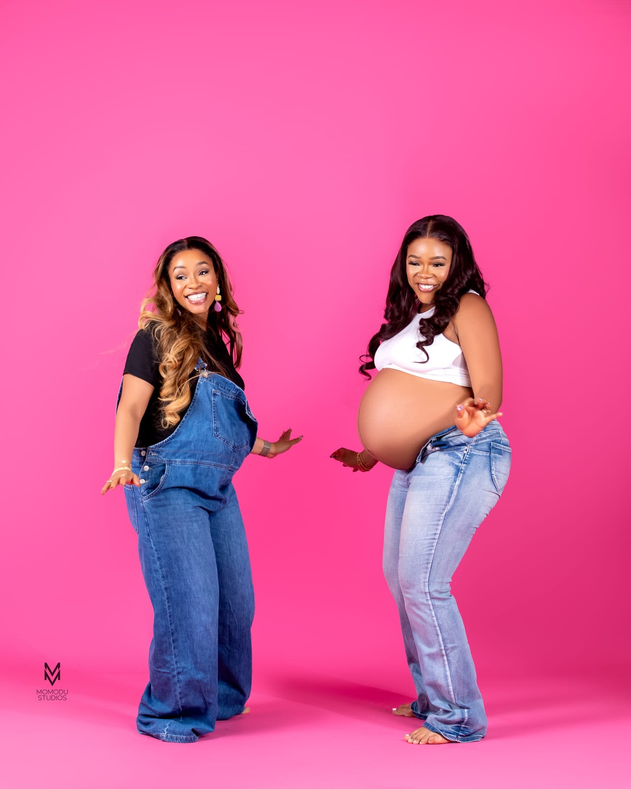 Two pregnant friends in denim and white tops smiling together on a pink set