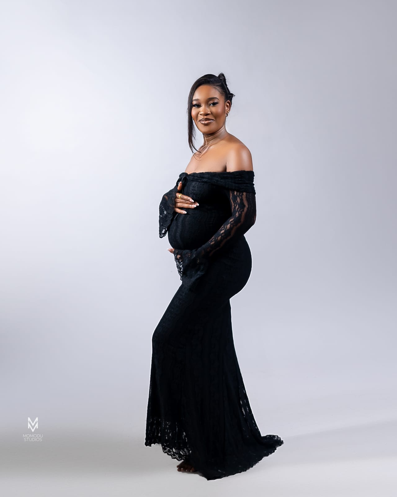 A pregnant woman in a black off-shoulder lace gown against a grey backdrop