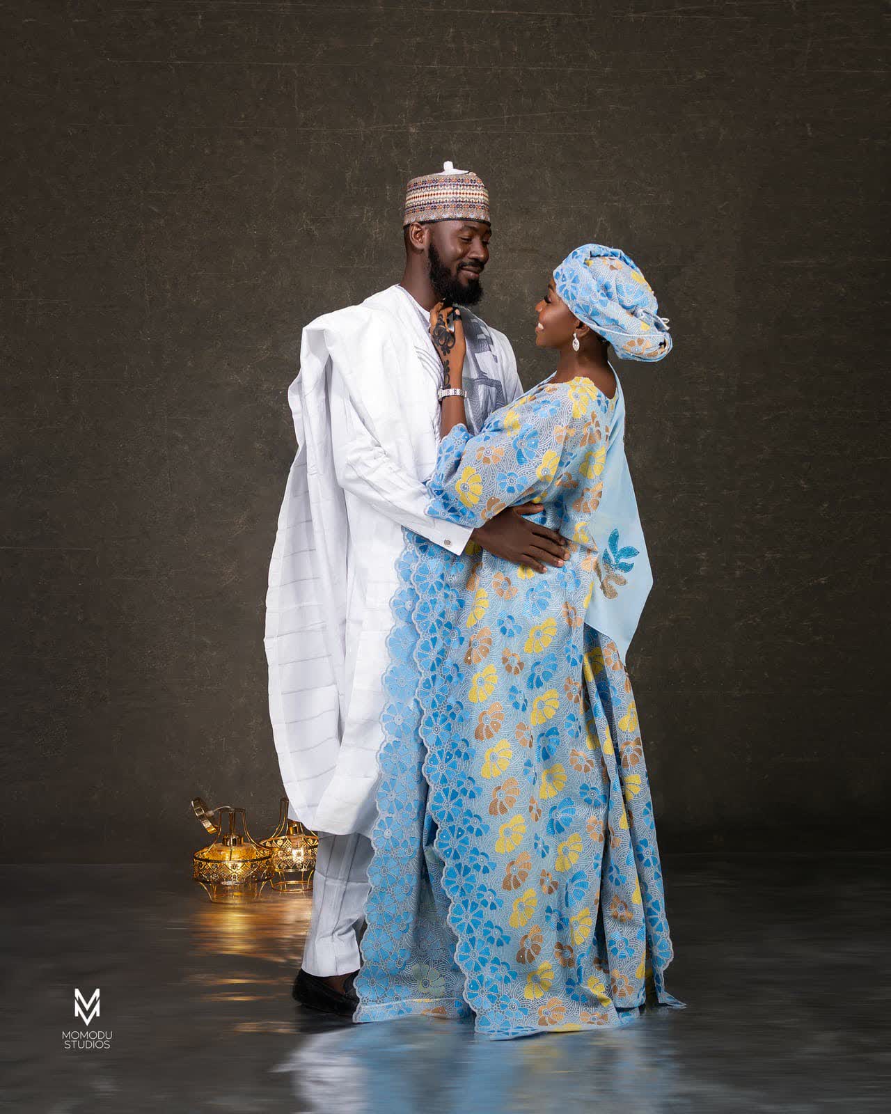 A couple in traditional attire, he in white agbada and cap, she in a blue gown and gele