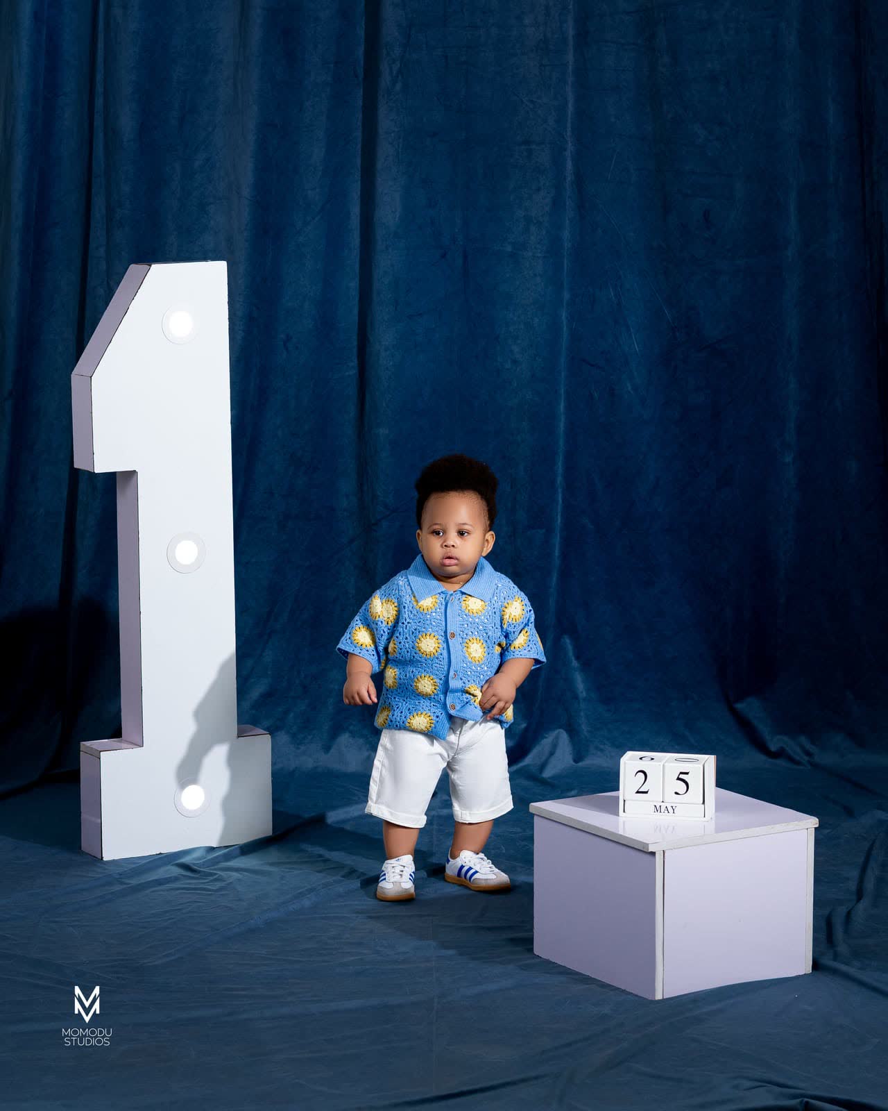 A toddler standing next to a large number one and a date block on a blue set