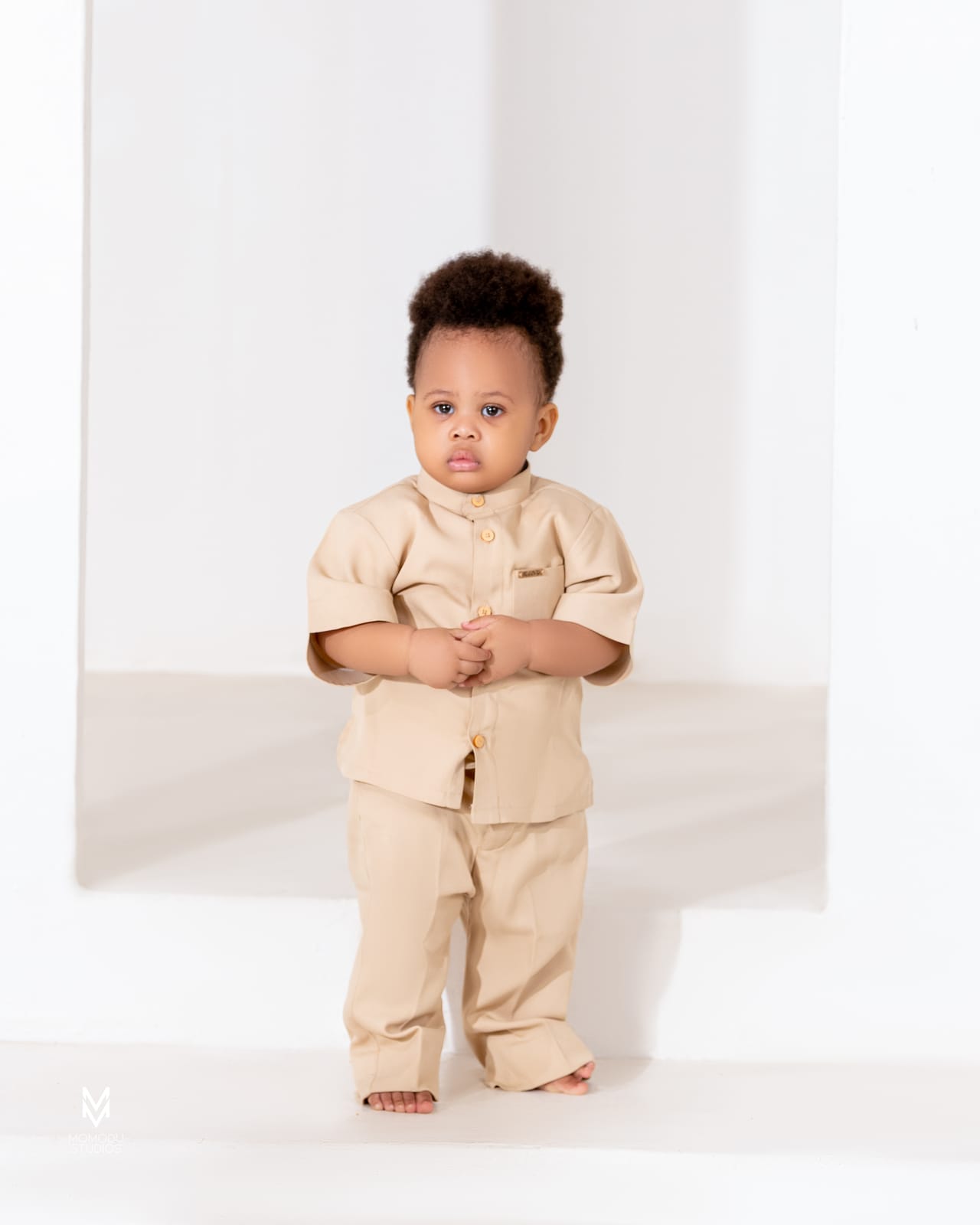 A toddler in a beige outfit standing against a white wall