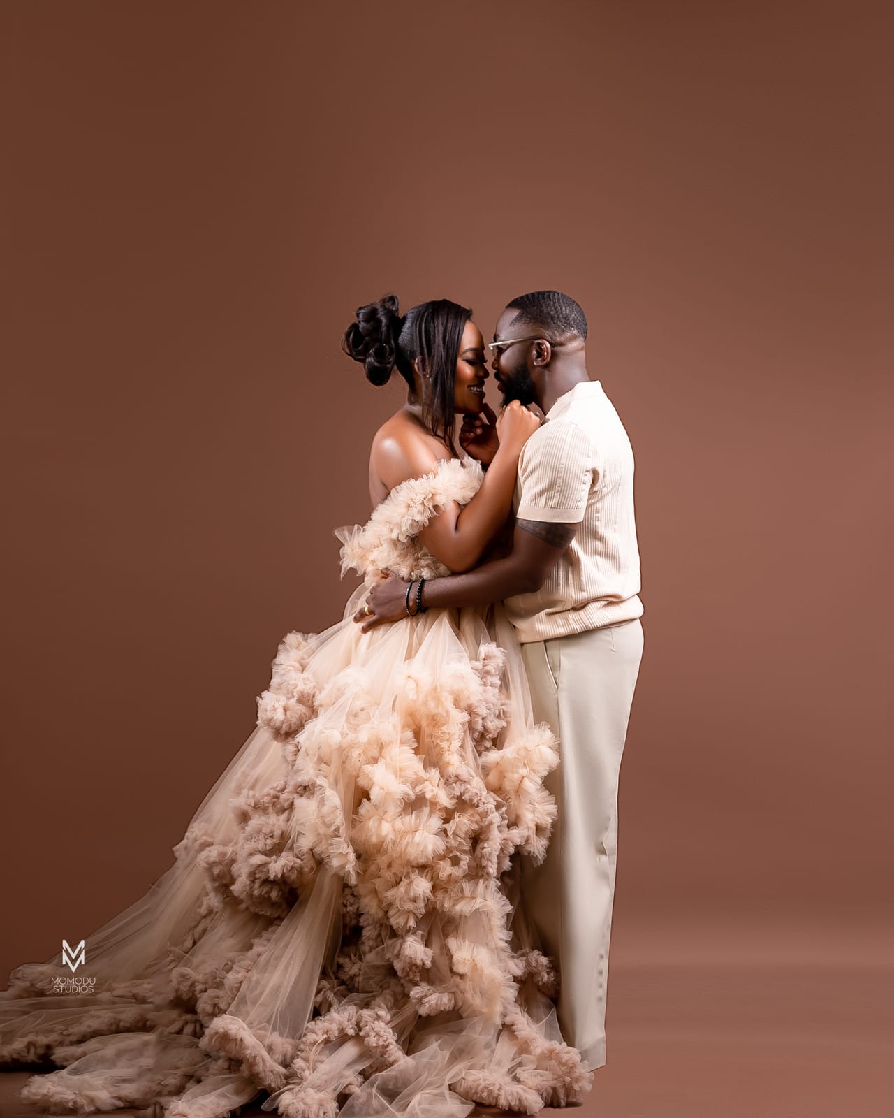 A couple kissing; she wears a voluminous ruffled blush gown, brown backdrop
