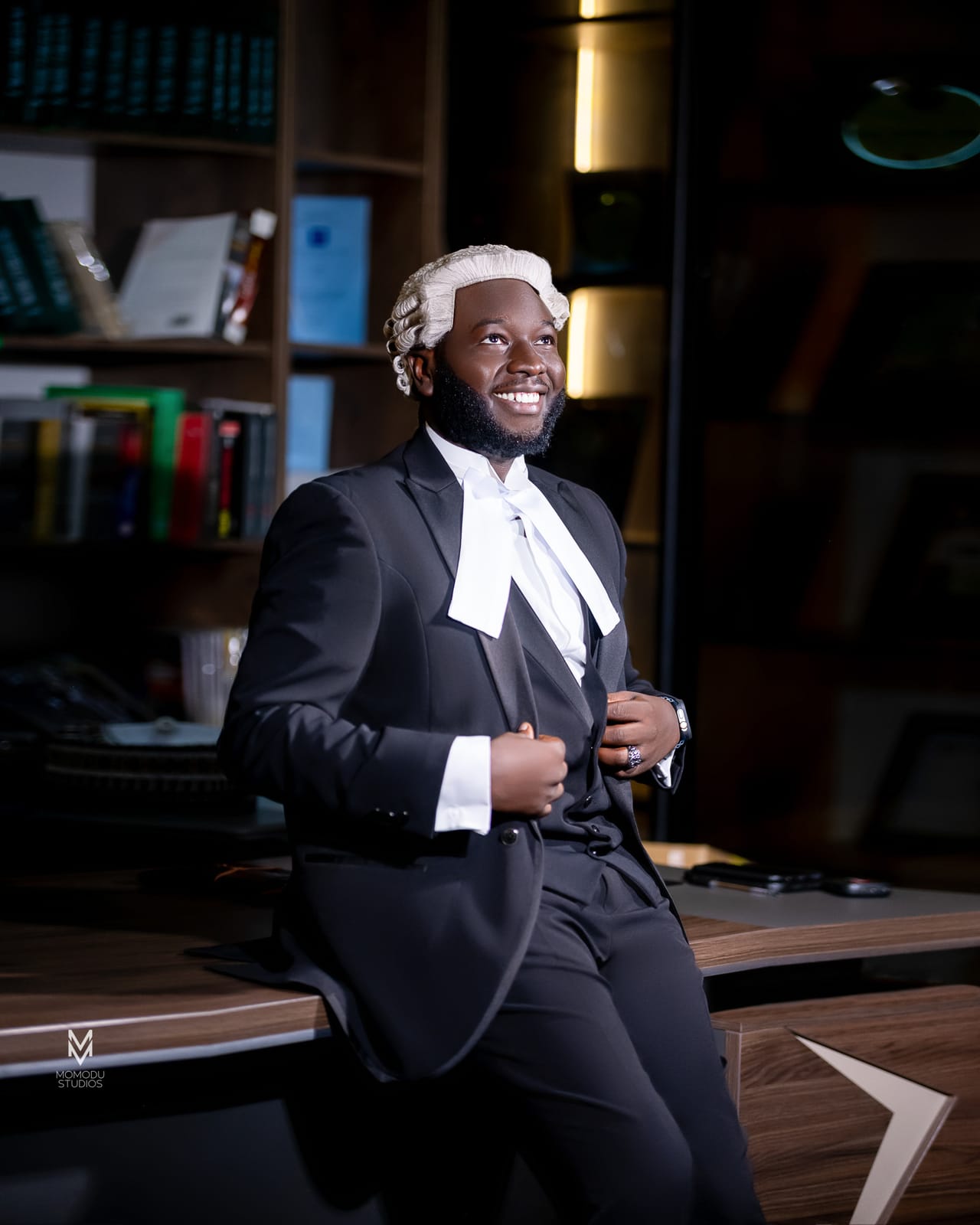 A smiling new lawyer in wig and suit leaning on a desk in an office