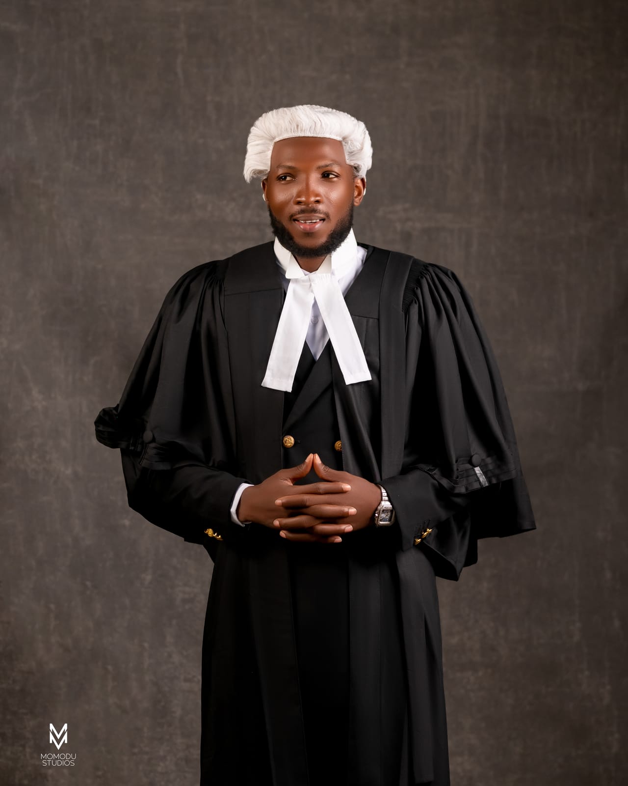 A new lawyer in wig and gown with hands clasped, grey backdrop