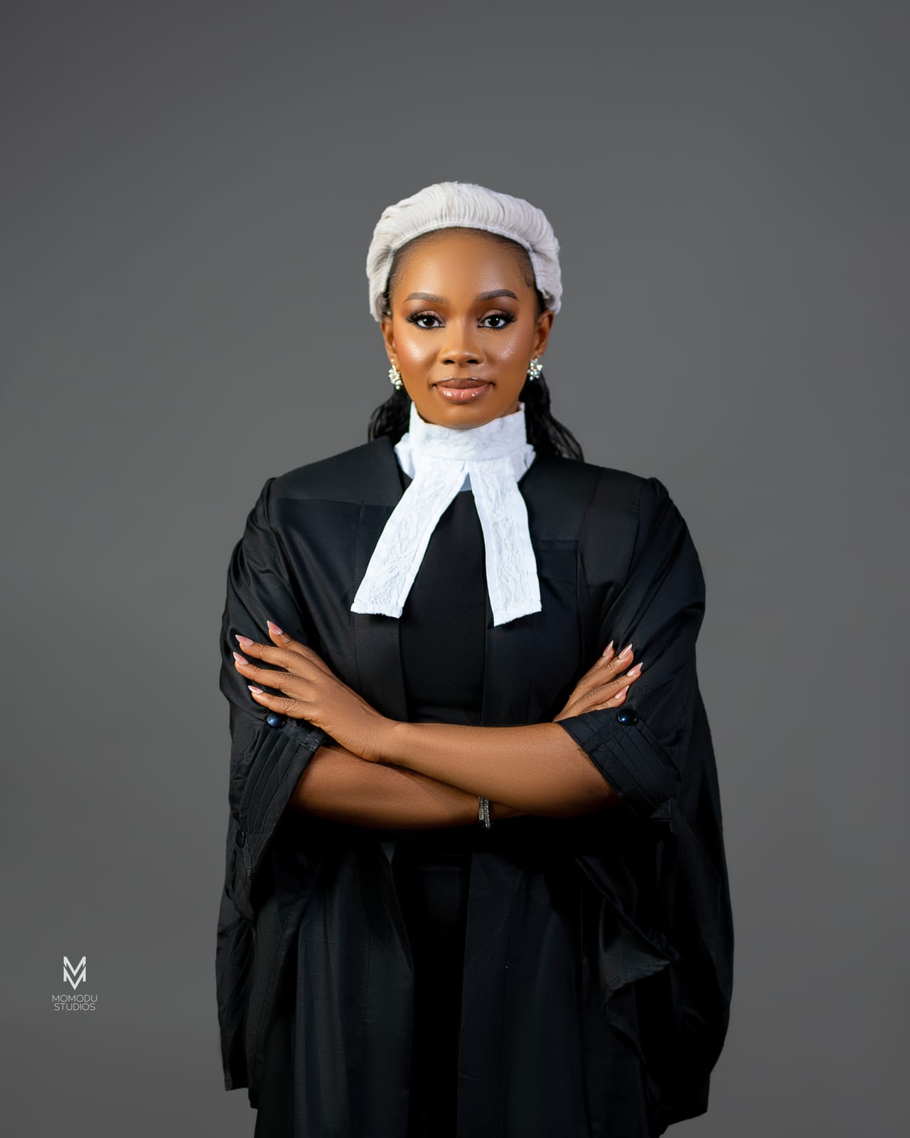 A woman in a barrister's wig and gown with her arms folded, grey backdrop