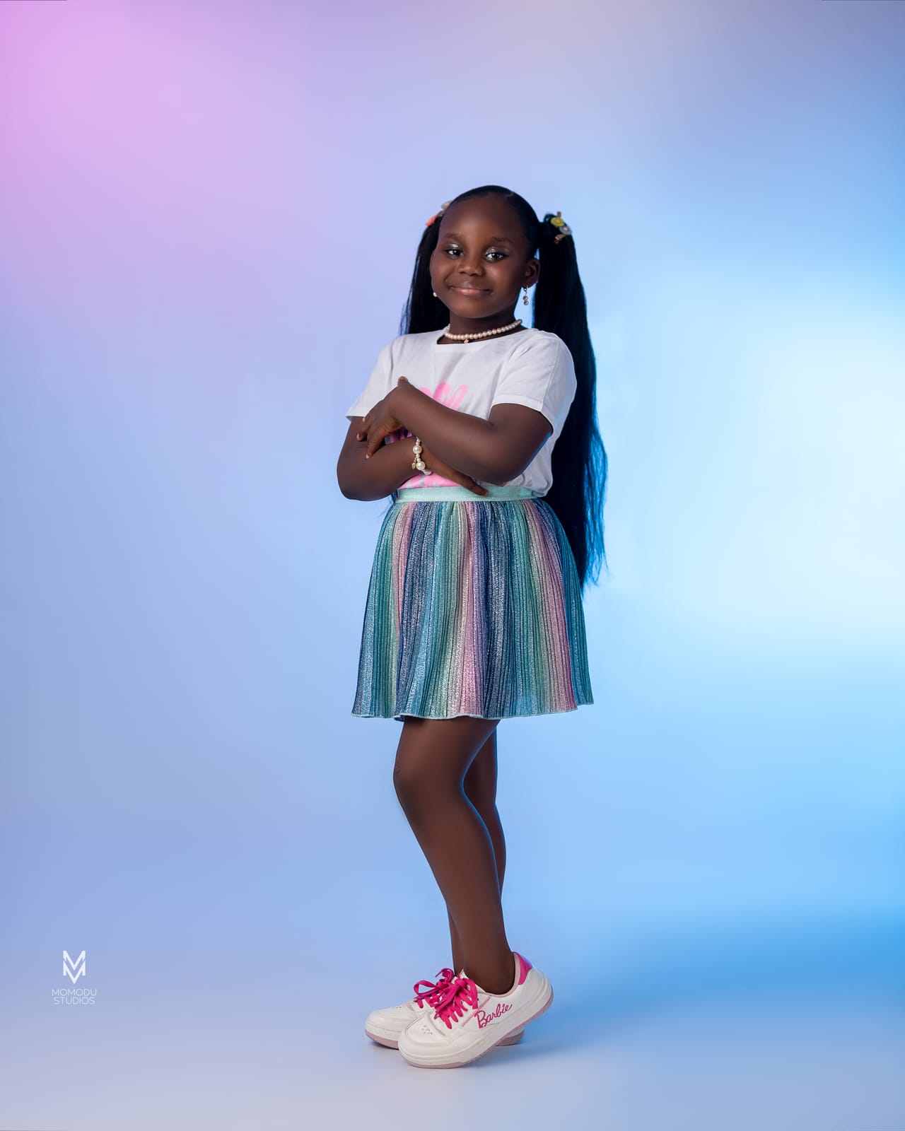 A girl in a white top and striped skirt posing with arms crossed, blue and pink backdrop