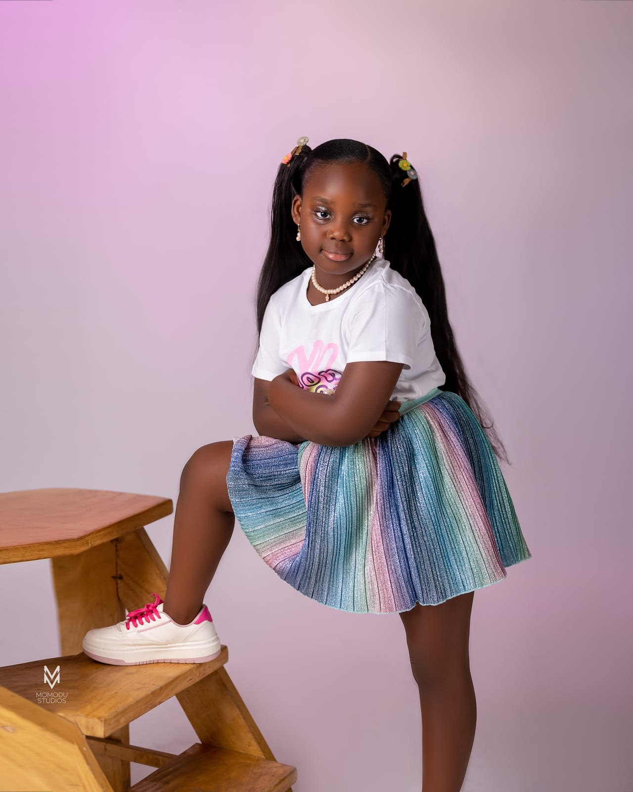 A smiling girl with one foot on a wooden stool, pastel backdrop