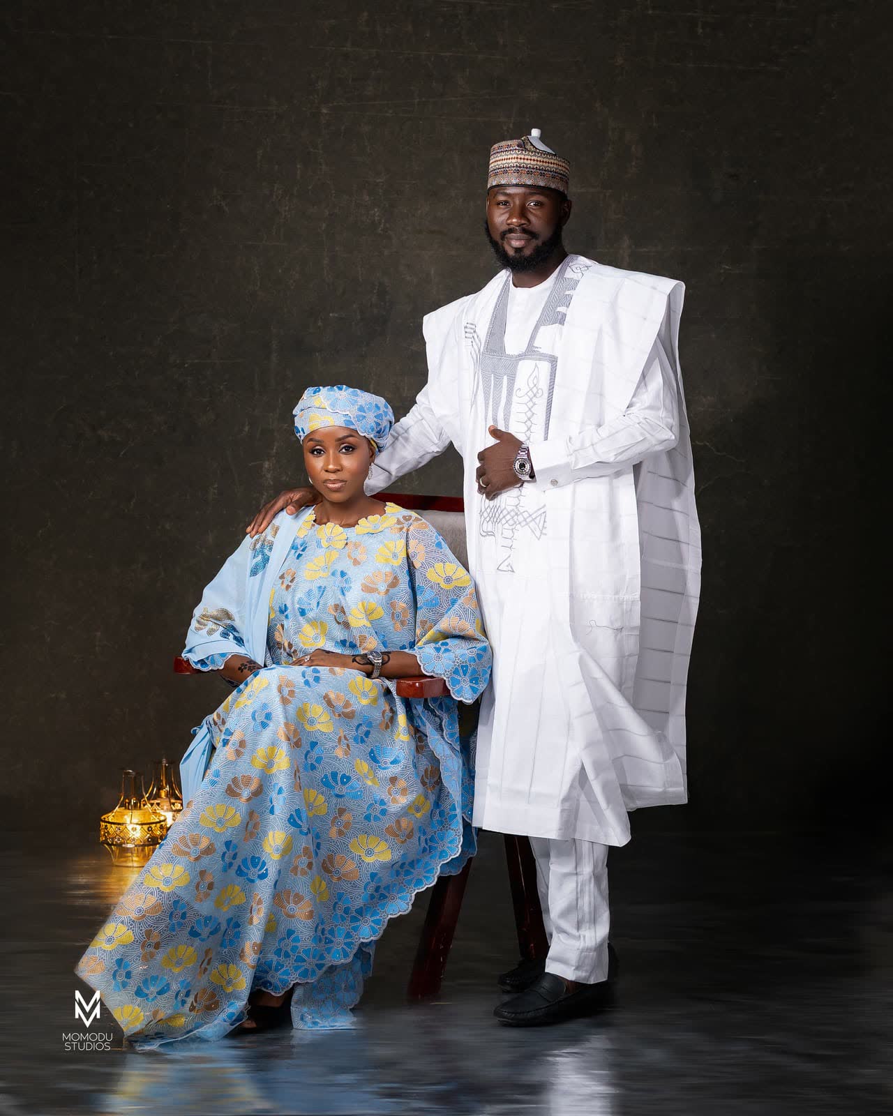 A woman seated in a blue patterned gown with a man in white agbada standing beside her