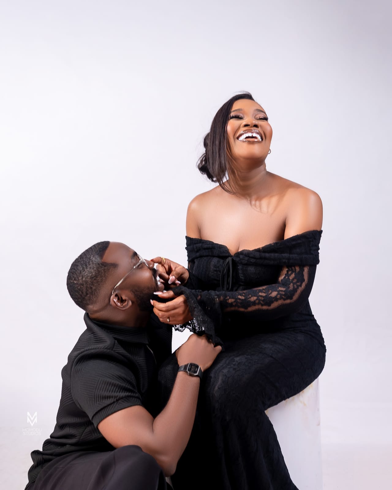 A man kneels to kiss the hand of a laughing woman in a black off-shoulder dress
