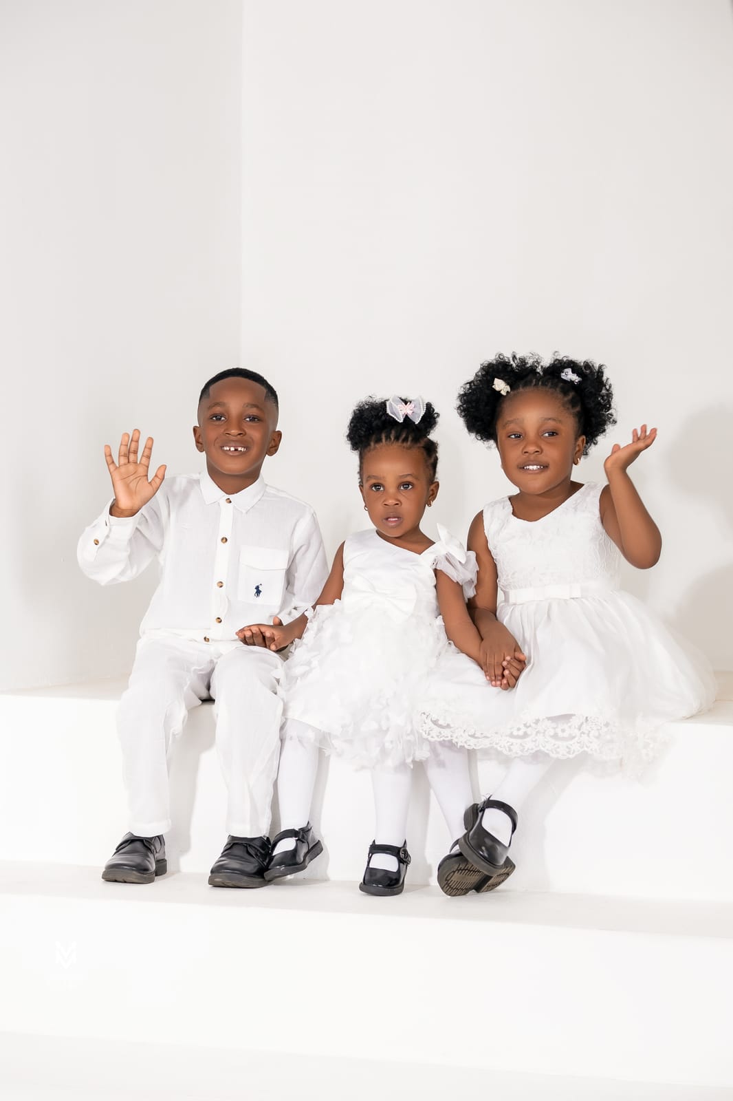 A boy and two little girls in white sitting on the studio floor and waving
