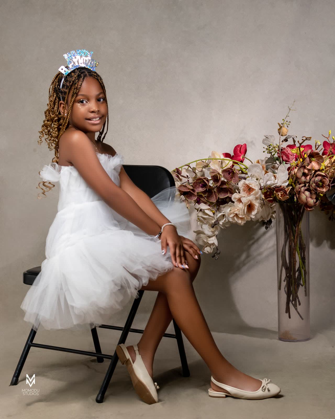 A girl in a white tulle dress seated beside a vase of dried flowers