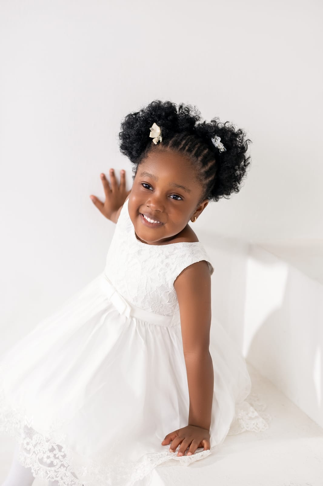 A smiling toddler with puffs in her hair and a white lace dress, waving